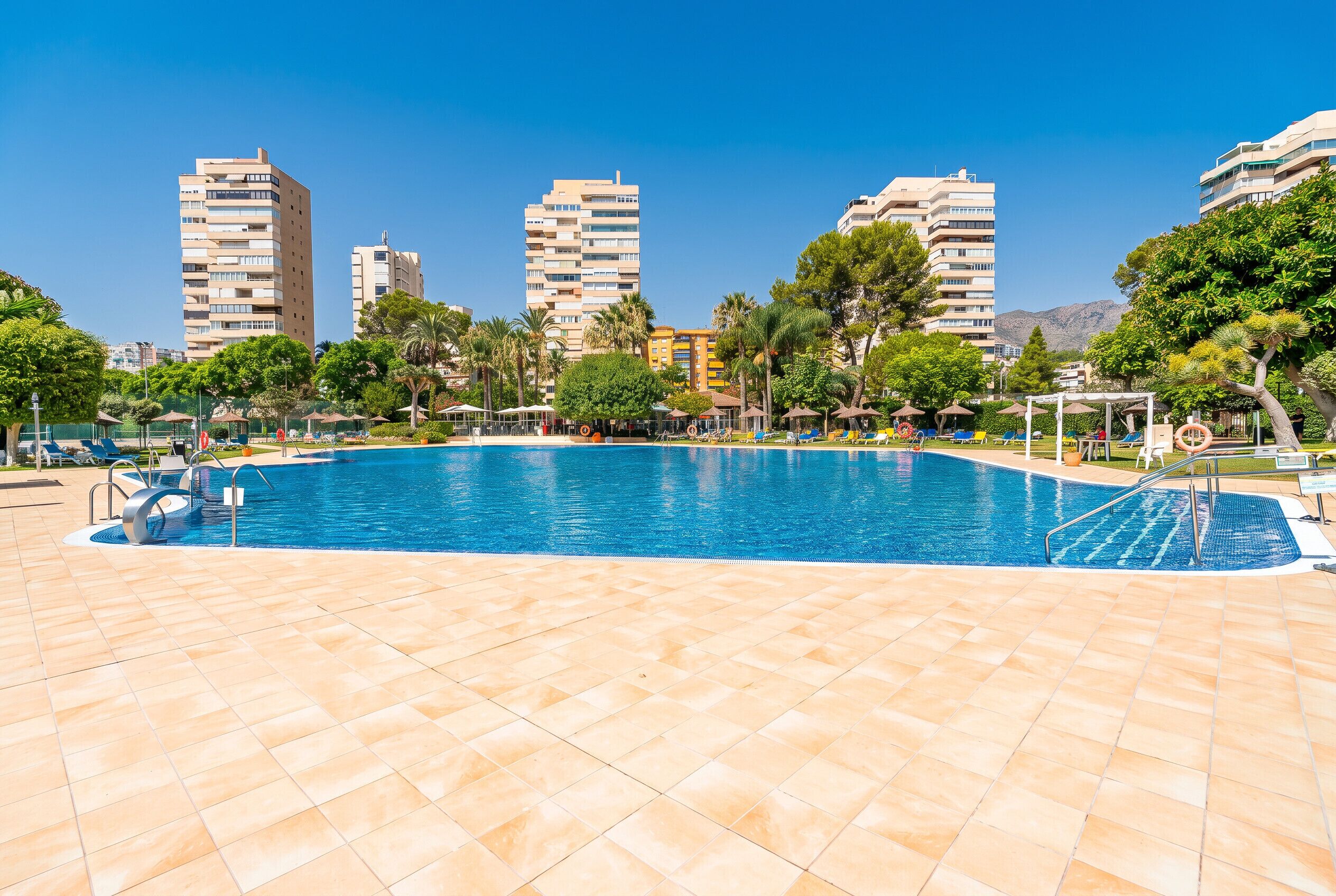 A swimming pool with tiled flooring, surrounded by palm trees and lounge chairs.