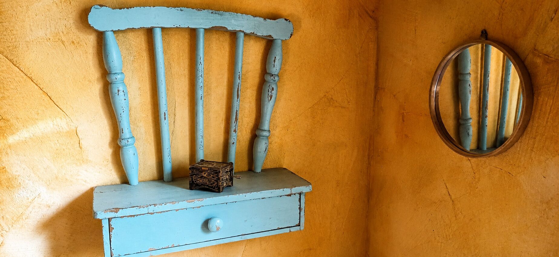 Una parete rustica con una mensola di legno azzurra, su cui sono posizionati un piccolo scatola e uno specchio.