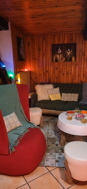 A cozy room with wooden walls, a red chair, a green sofa, and a round table with snacks and drinks.