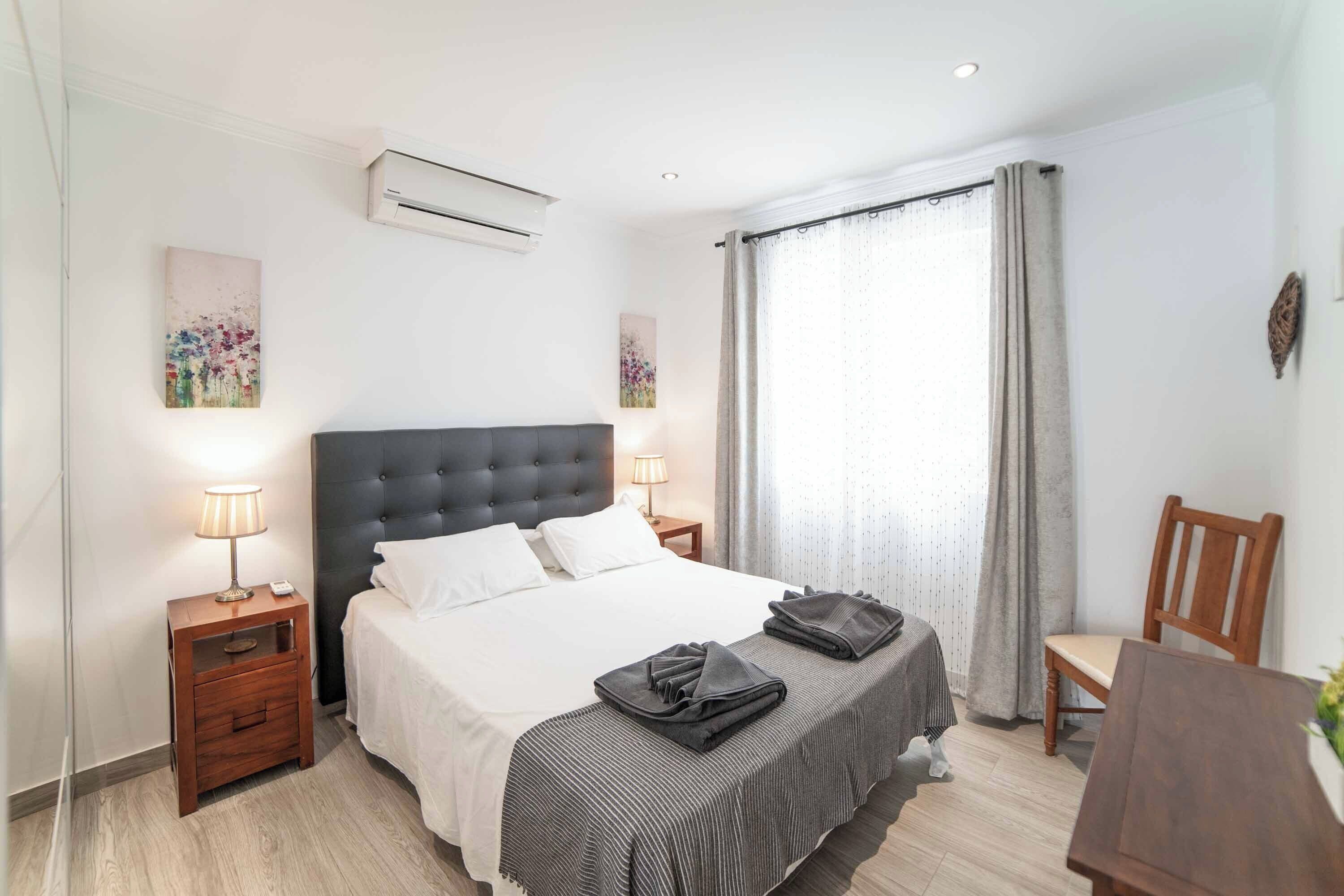 Une chambre d’hôtel avec un lit, des lampes de chevet, une petite table en bois, une chaise et une fenêtre avec des rideaux.