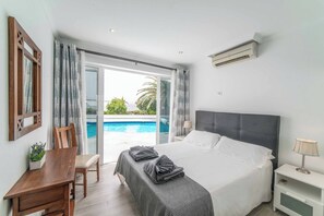 A bedroom with a bed, a desk, a chair, a nightstand, a lamp, and a view of a swimming pool. - Villa Botanica - Four Bedroom Villa, Sleeps 8 (Playa Blanca)