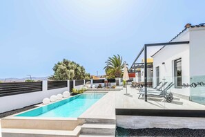 Een modern buitengebied met een zwembad in helderblauw water, een terras met ligbedden, en een gebouw met een witte gevel en een bord.