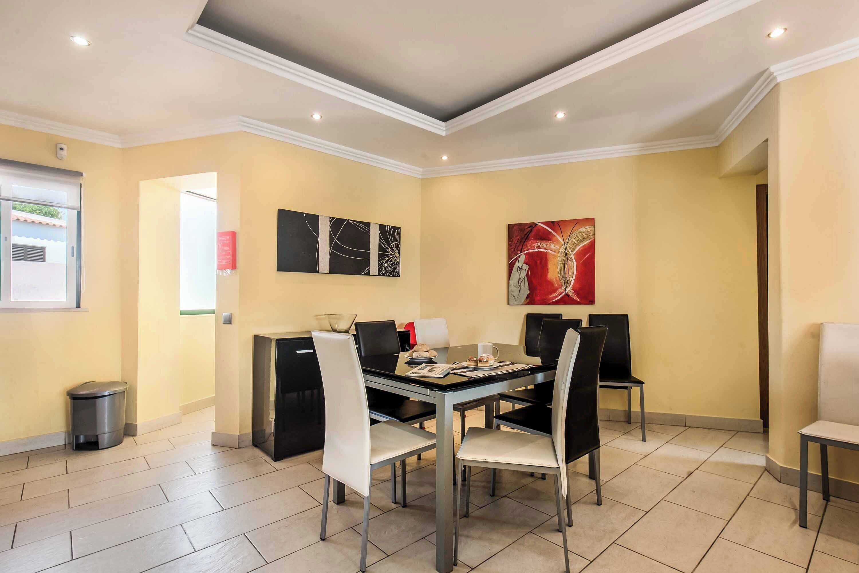 Uma sala de jantar com uma mesa posta para quatro pessoas, rodeada por cadeiras, com paredes em tons de bege e piso ladrilhado.