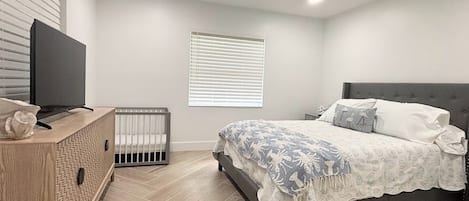 Une chambre à coucher avec un lit, un téléviseur posé sur un meuble en bois, un berceau et une fenêtre avec des stores.