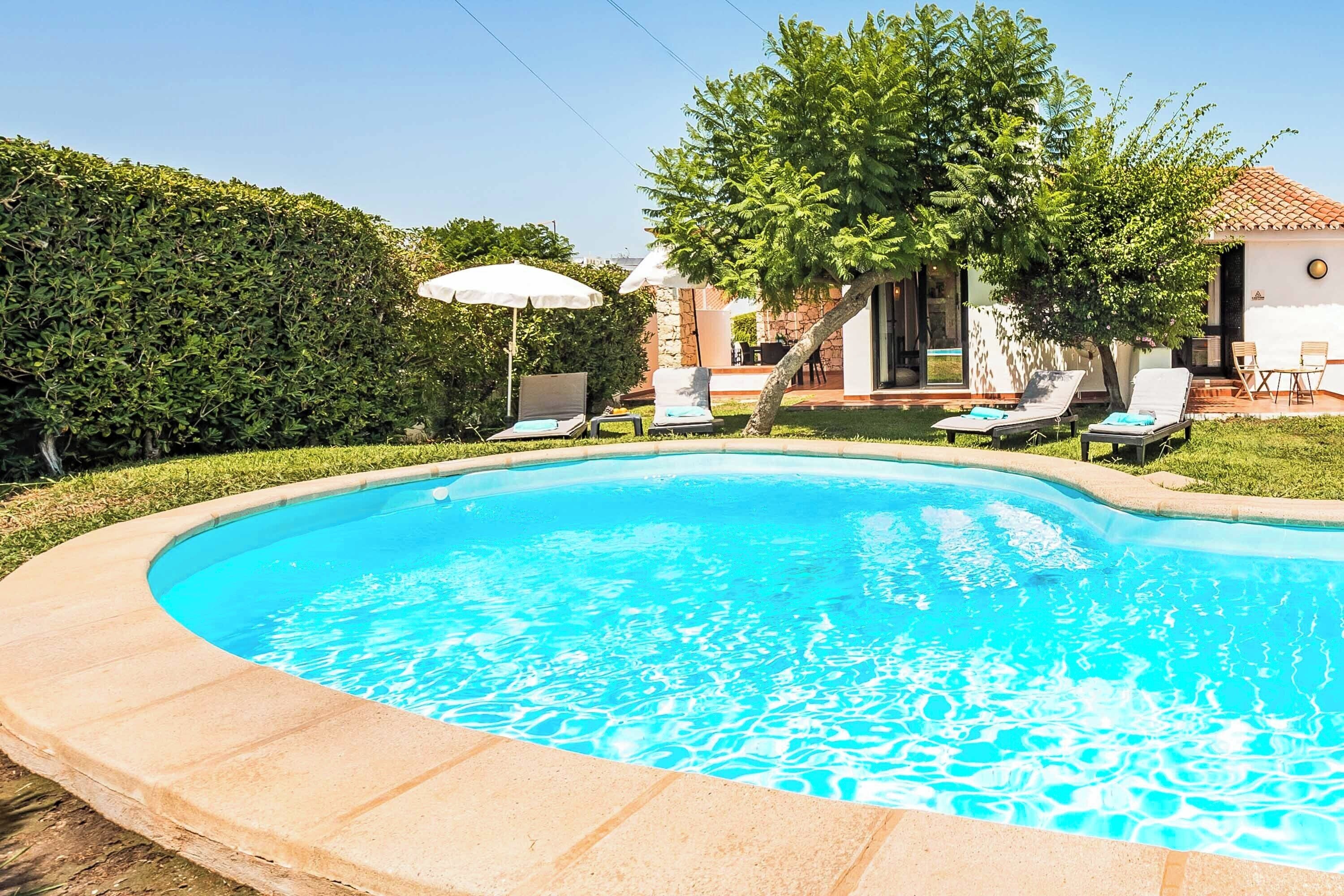 A swimming pool with lounge chairs and umbrellas, surrounded by greenery and a building.
