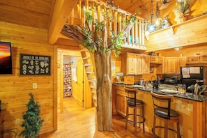 A wooden cabin interior with a central tree, a kitchen area with bar stools, and a sign listing animals of the woodlands. - Creekview Cabin with Treehouse Loft (Sevierville)