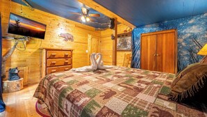 Um quarto de cabana aconchegante com cama, paredes de madeira, ventilador de teto, televisão e uma estátua de urso.