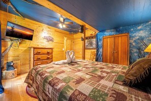 Ein gemütliches Blockhausszimmer mit Bett, Holzwänden, Deckenventilator, Fernseher und einer Bärenstatue.