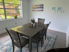 A dining area with a table set for four, surrounded by chairs, a window with a view of greenery, and a wall with framed artwork. - Beautifull updated modern single 2-bedroom single home close to beaches & shops (Holiday)