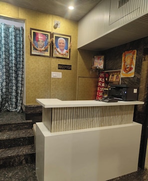 A reception desk with a sign above it, a staircase leading to an upper level, and a wall with framed pictures and a decorative garland. - Hotel Sangam Rajhans (Jaipur)