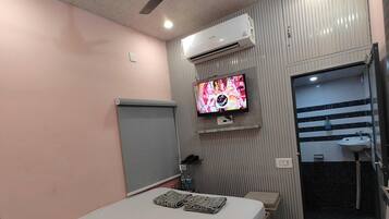 A hotel room with a bed, a television mounted on the wall, an air conditioning unit, a ceiling fan, and a bathroom with a sink and shower.