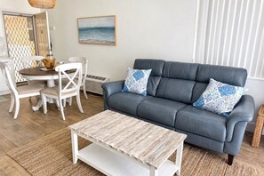 Ein Wohnzimmer mit einem grauen Sofa, zwei gemusterten Kissen, einem Holztisch, weißen Stühlen und einem Esstisch.