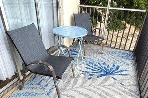 Ein Balkon mit zwei Stühlen und einem kleinen Tisch, einem gemusterten Boden und Blick auf Bäume durch ein Metallgeländer.