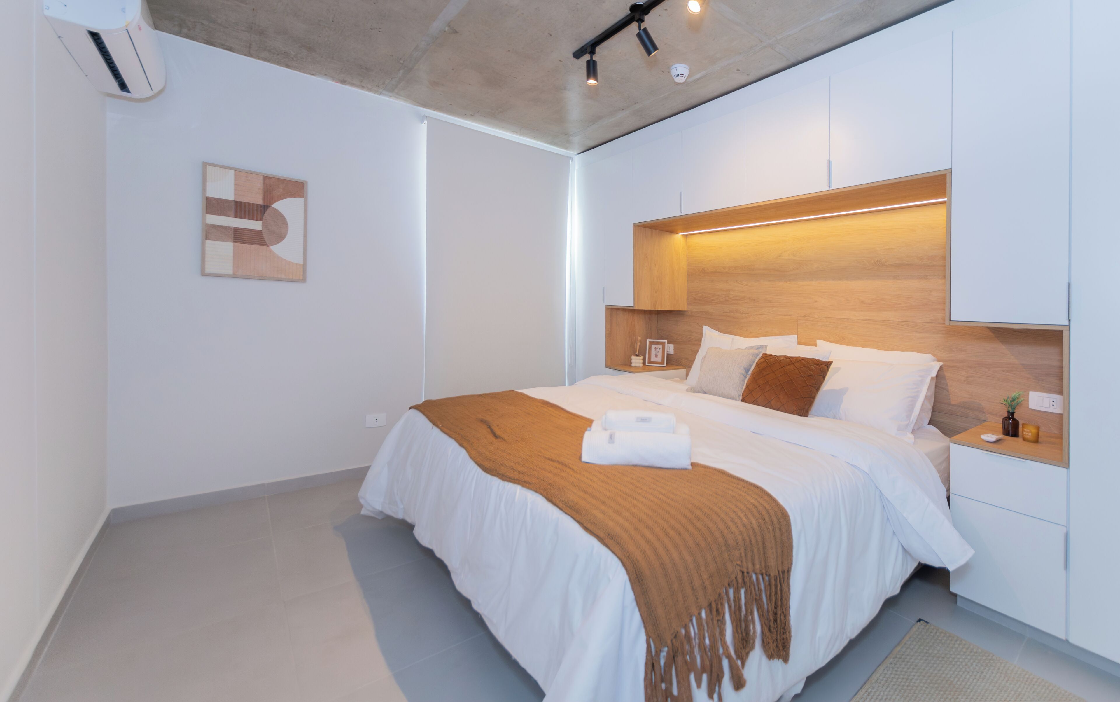 A modern bedroom with a large bed, a headboard with integrated lighting, a bedside table with a lamp, and a wall-mounted air conditioning unit.