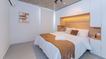 A modern bedroom with a large bed, a headboard with integrated lighting, a bedside table with a lamp, and a wall-mounted air conditioning unit.