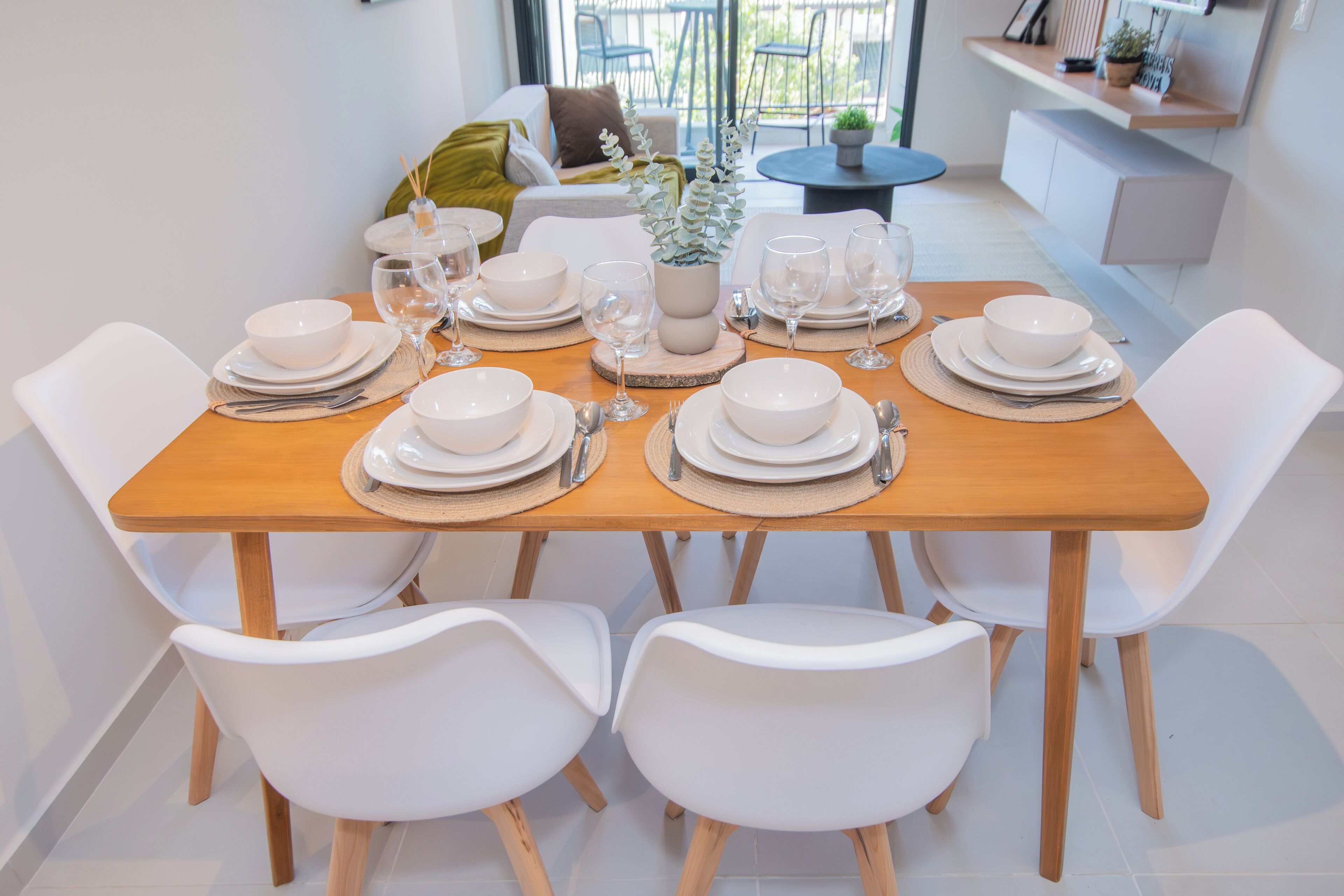 A dining table set with white plates, bowls, and cutlery, surrounded by white chairs in a modern interior.