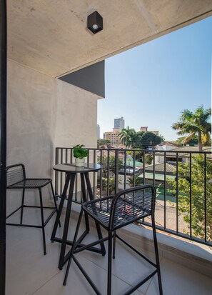Ein Balkon mit Tisch und zwei Stühlen, einem Blumentopf und Blick auf Gebäude und Grünflächen.