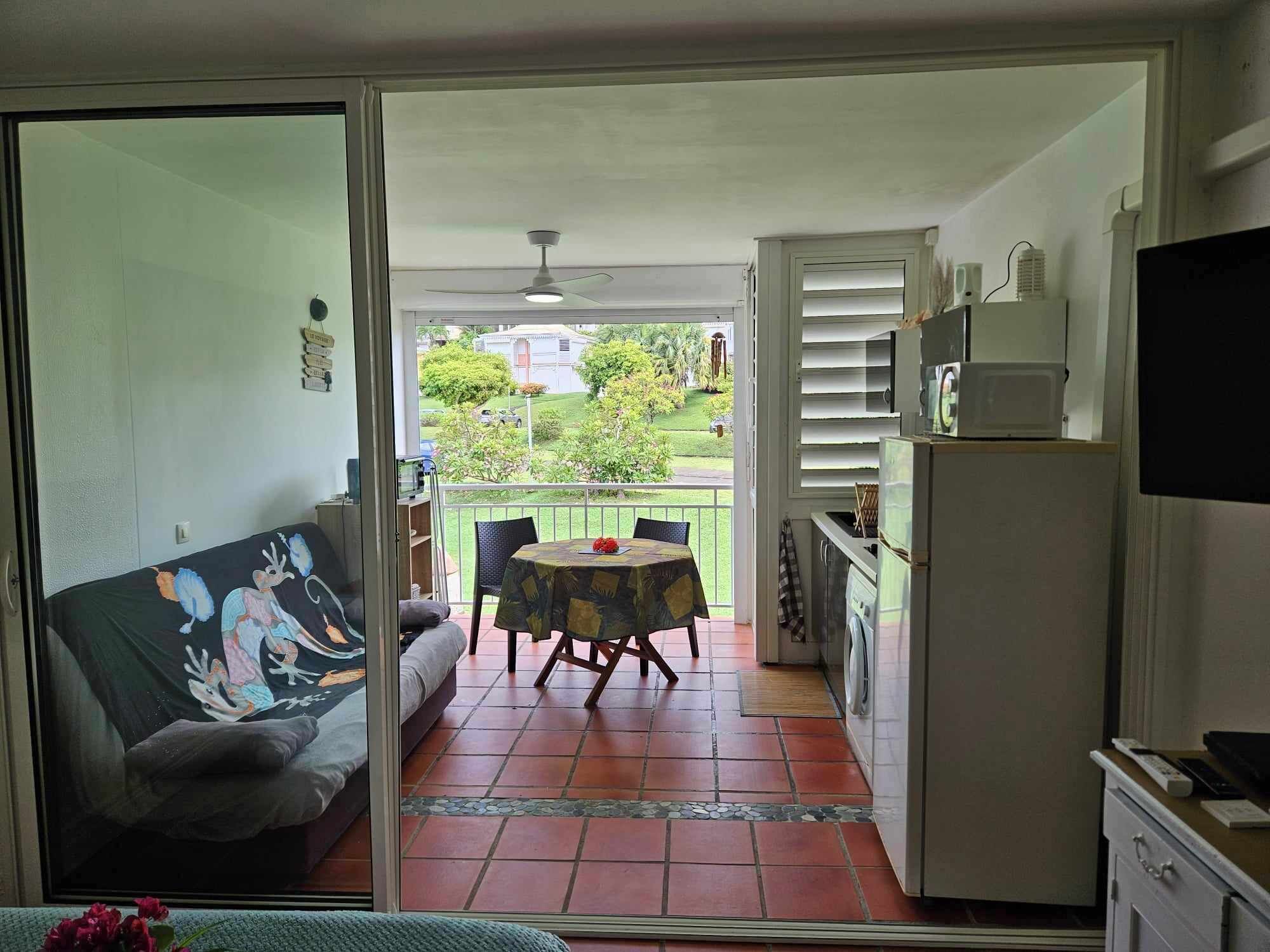A room with a sliding glass door leading to a balcony, a dining table with chairs, a sofa, a washing machine, and a television.
