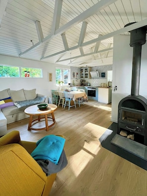 A spacious living area with a sofa, a round wooden table, and a kitchen with a built-in wood stove. - Stay in" Sandersvig delight" filled with colours (Haderslev)