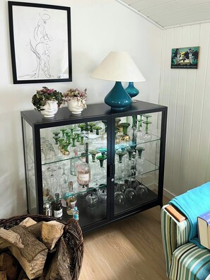 Ein Raum mit einer gläsernen Vitrine, in der verschiedenes Glasbesteck und Flaschen ausgestellt sind, einer blauen Lampe auf einer Konsole und einem gerahmten Kunstwerk an der Wand.