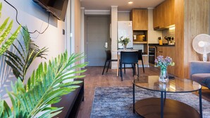 A modern living room with a flat-screen TV mounted on the wall, a dining area with a table and chairs, and a kitchen with wooden cabinets and a built-in oven. - Pedro Valdivia Apartments by Andes STR (Providencia)