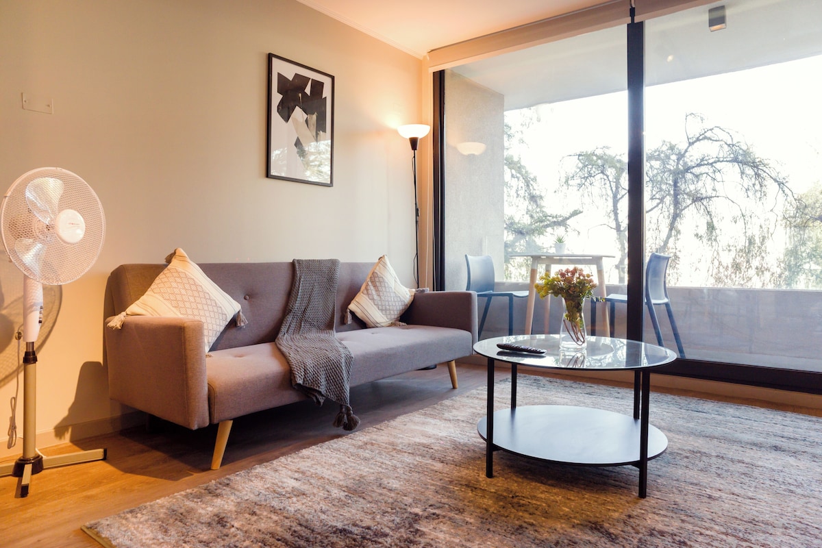 A modern living room with a sofa, a standing fan, a glass coffee table, and a balcony with outdoor furniture.