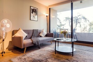 Een moderne woonkamer met een bank, een staande ventilator, een glazen salontafel en een balkon met tuinmeubilair.