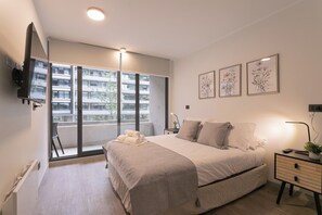 A modern bedroom with a large bed, a bedside table, a TV mounted on the wall, and a balcony with a view of a building. - Pedro Valdivia Apartments by Andes STR (Providencia)