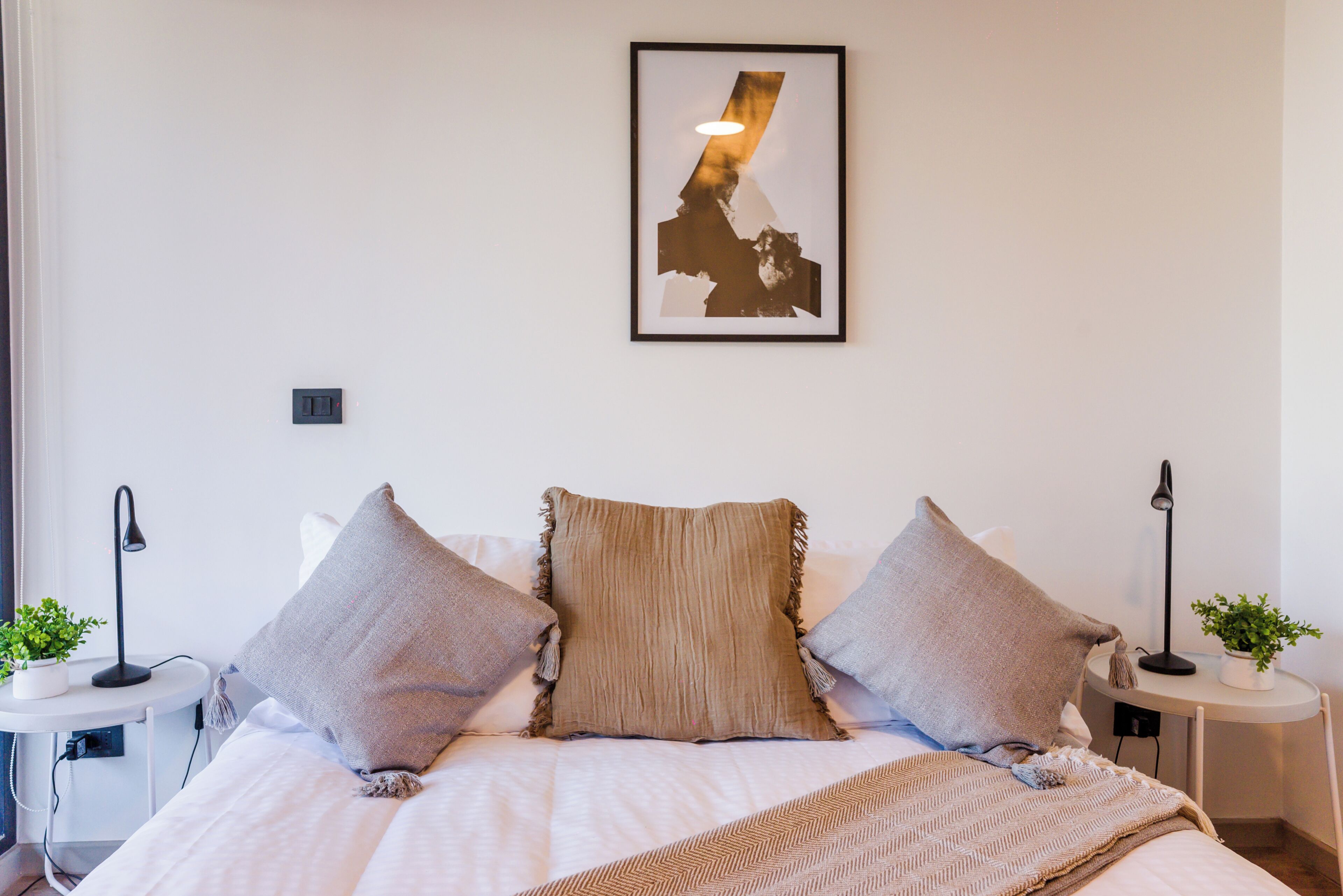 Een net opgemaakt bed met drie kussens, een wandlamp en een ingelijst kunstwerk erboven.