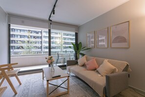 Een moderne woonkamer met een bank, salontafel en stoelen. Er is een groot raam met uitzicht op een gebouw en een plant.