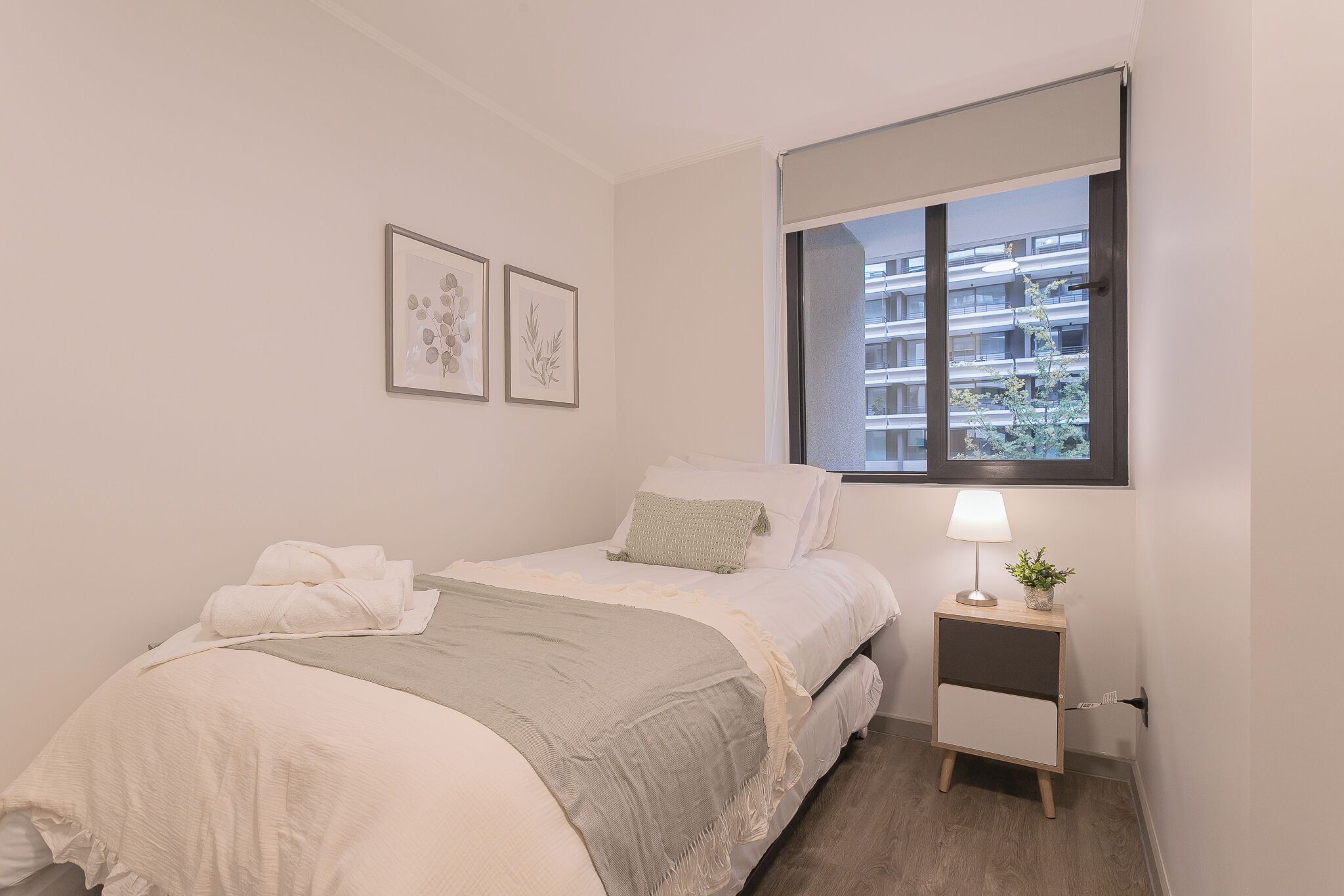 Een minimalistische slaapkamer met een bed, een nachtkastje met een lamp, en twee ingelijste botanische prints aan de muur.