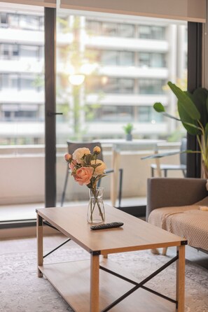 A modern living room with a wooden coffee table, a vase with flowers, a remote control, and a view of a cityscape through large windows. - Pedro Valdivia Apartments by Andes STR (Providencia)