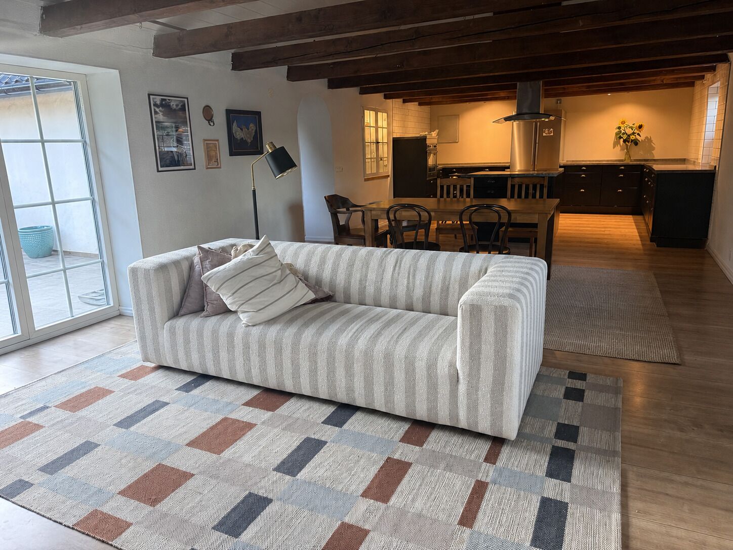 Una sala de estar con sofá, alfombra estampada, vigas de madera, comedor y cocina.