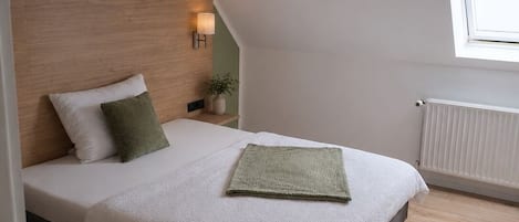 A bedroom with a sloped ceiling, a bed with white bedding, a bedside table with a lamp, and a window.