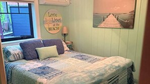 Um quarto com uma cama, um ventilador de teto, uma janela e um quadro na parede.