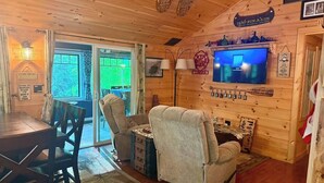 Interior de cabana com mesa de jantar, duas poltronas, televisão e lareira.
