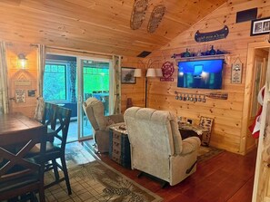 Ein Blockhaus-Interieur mit Esstisch, zwei Sesseln, einem Fernseher und einem Kamin.