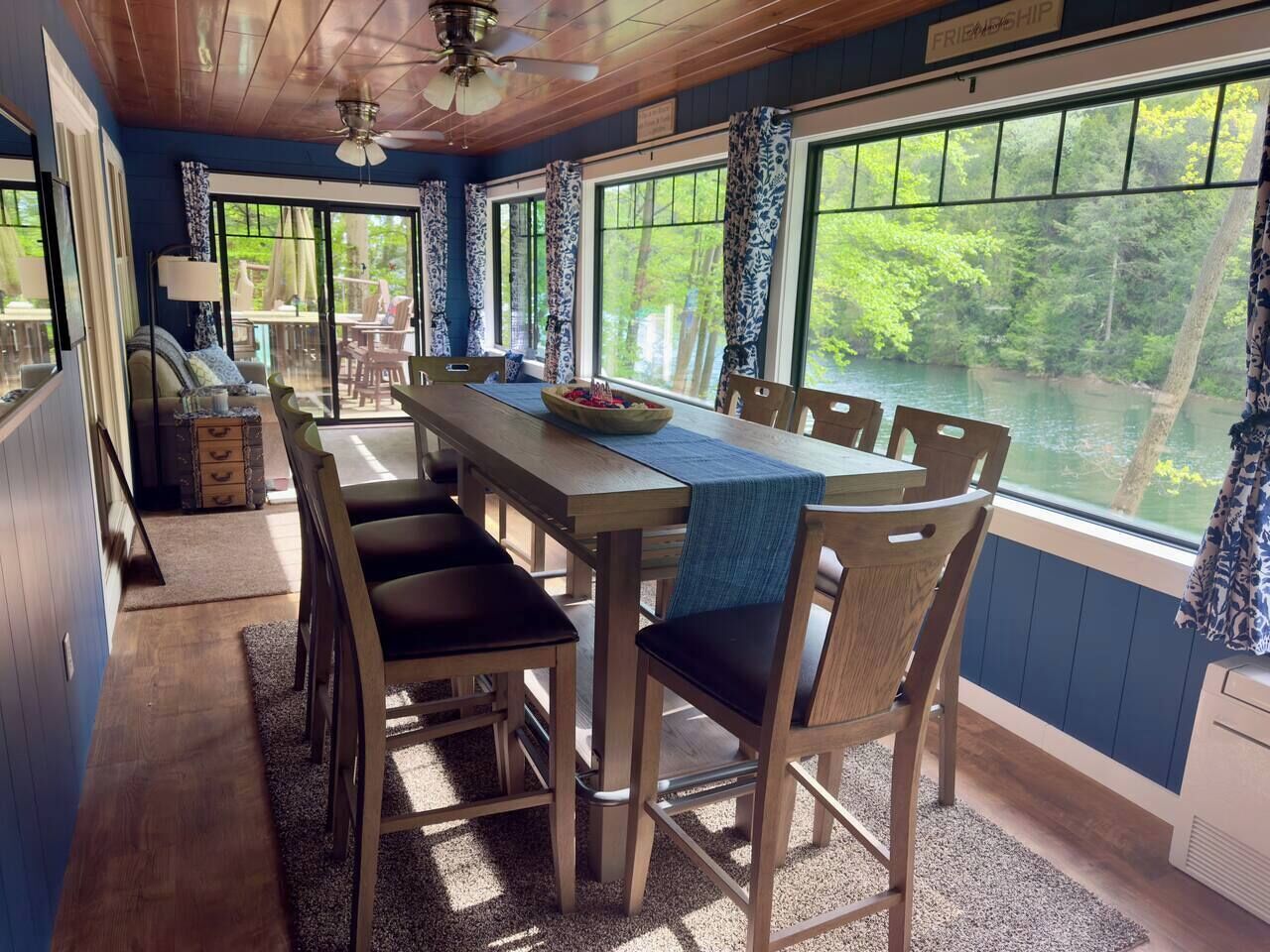 A dining area with a long table and chairs, large windows with curtains, and a view of a natural landscape.