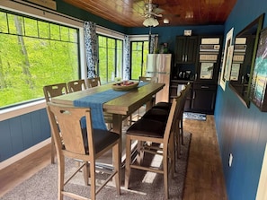 A dining area with a wooden table and chairs, a refrigerator, and a view of trees through large windows. - Peaceful Lakefront Retreat,  Fish, Kayak, Unplug 18 Min from Middlebury (Leicester)