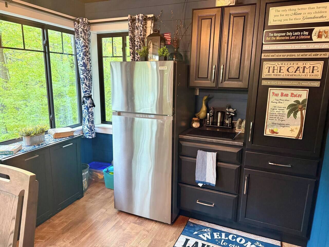 Una cocina con un refrigerador de acero inoxidable, armarios de madera, un ventanal con vista a follaje y un tapete de bienvenida.