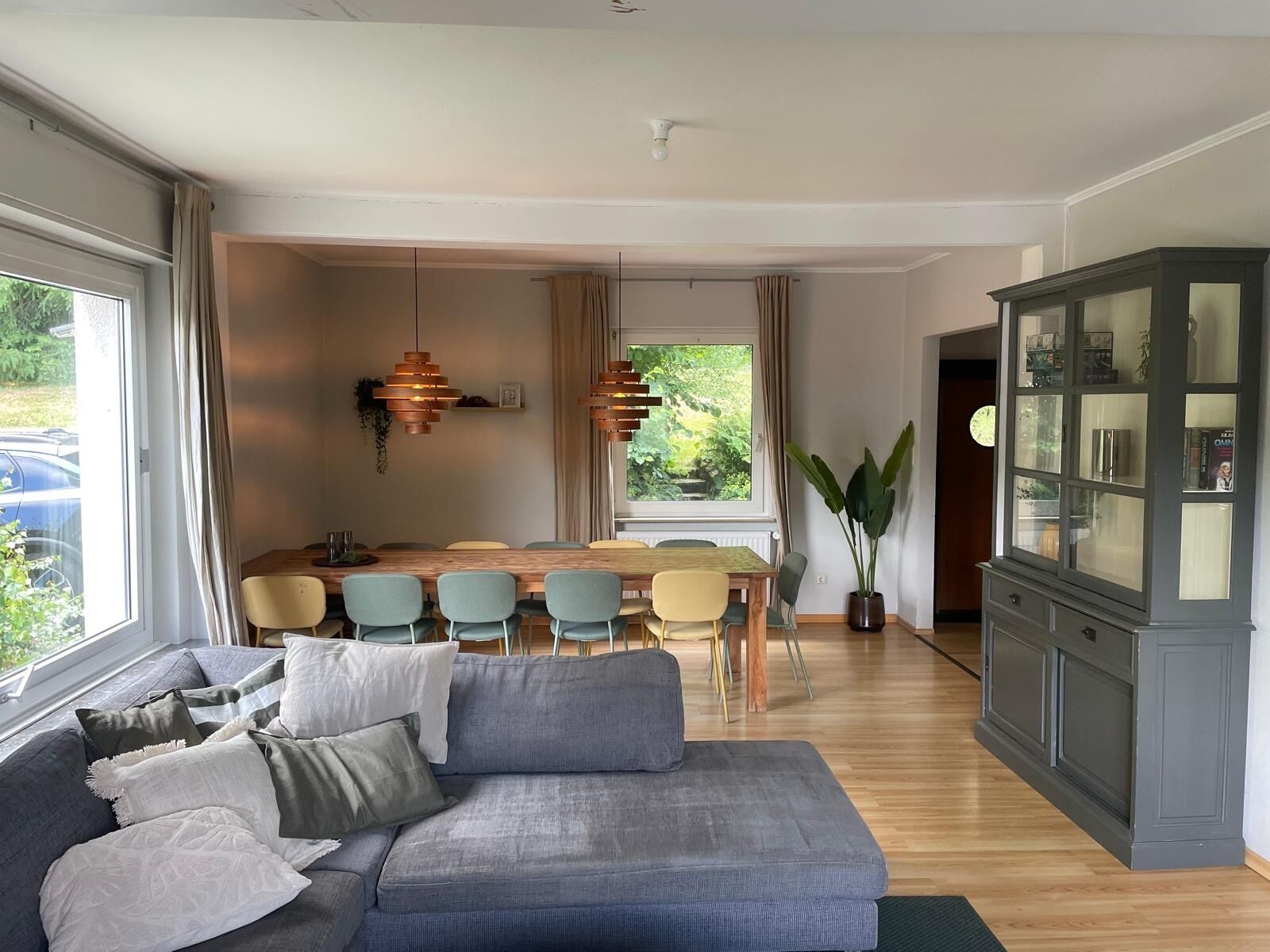 A modern living room with a grey sofa, dining area with a wooden table, and a cabinet with glass doors.