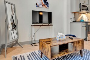 Ein modernes Wohnzimmer mit Couchtisch aus Holz, einem Flachbildfernseher, einem Gemälde und einer Küchenzeile im Hintergrund.