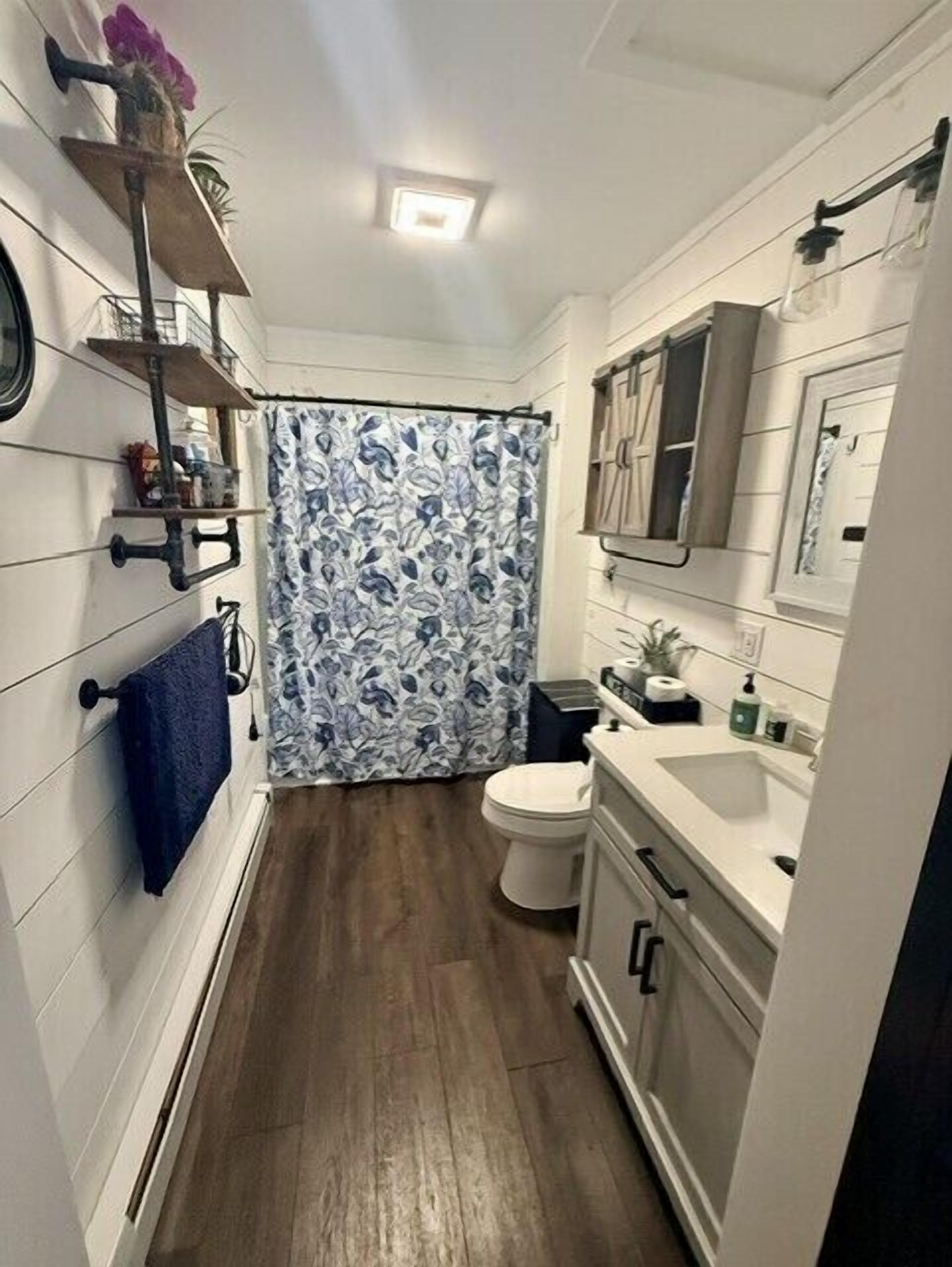 Ein schmales Badezimmer mit Toilette, Waschbecken und Spiegel. Ein Blumenmuster ziert die Duschvorhänge.