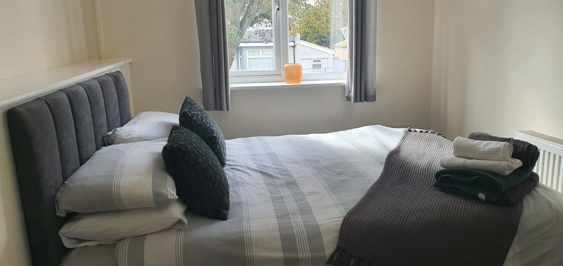 A neatly made bed with pillows and a folded blanket, a window with curtains, and a radiator on the wall.