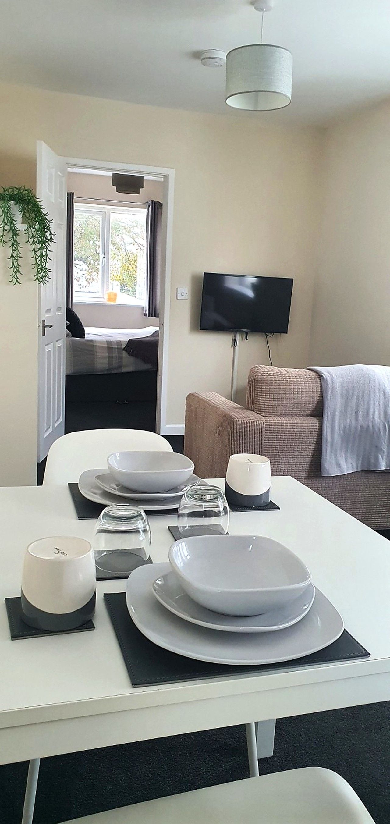 A dining area with a table set for two, a television mounted on the wall, and a sofa in the background.