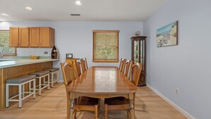 A dining room with a long wooden table and chairs, a kitchen with wooden cabinets, and a window with blinds. - SH30: Southern Shores 30 (Southern Shores)