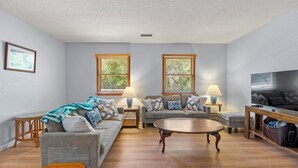 A living room with a sofa, a coffee table, a TV, and two windows with blinds. - SH30: Southern Shores 30 (Southern Shores)
