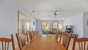 A spacious dining room with a long wooden table and chairs, a living area with a sofa and television, and large windows with blinds. - SH30: Southern Shores 30 (Southern Shores)