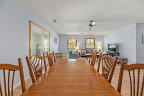 Ein geräumiger Essbereich mit einem langen Holztisch und Stühlen, eine Wohnarea mit Sofa und Fernseher sowie große Fenster mit Jalousien.