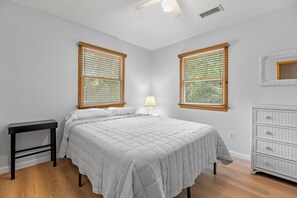 Ein Schlafzimmer mit Bett, Nachttisch, Lampe, Kommode und zwei Fenstern mit Jalousien.